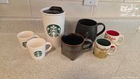 Photo showing all seven Starbucks mugs and cups arranged on counter: two small white demitasse cups, one large white travel tumbler with Starbucks logo, one black Reserve Roastery NY mug, one Hangzhou mug, one China mug with red interior.