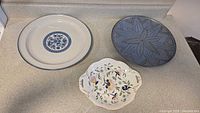Three serving trays on a countertop: a small ornate floral plate with gold accents, a round blue and white plate with a floral pattern, and a blue pottery dish with a geometric design.