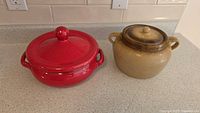 Two stoneware casserole dishes with lids placed on kitchen countertop, showing the red and tan/brown dishes side by side.