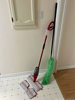 Photo of the complete mop and duster set including the Vileda floor spray mop standing upright, two mop pads on the floor, and the green extendable duster leaning against the wall.