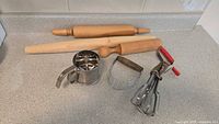 Lot of vintage baking tools on counter, including two wooden rolling pins, metal pastry blender, hand-cranked flour sifter, and hand egg beater with red handles.