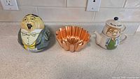 Three items displayed on countertop: ceramic teapot with 'Tea for Father' illustration, copper jelly mold with scalloped edges, vintage tin with character face in tuxedo.