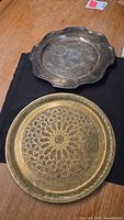 Two vintage metal decorative plates displayed on dark cloth. Top is scalloped metal plate with wear and scratches. Bottom is large copper tray with complex geometric engraved design.