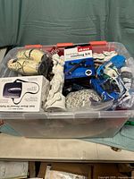 View of clear plastic storage tote containing a variety of items including a boxed cooling kit, white classical figurine, and various wrapped objects.