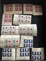 Top view of multiple blocks of unused Canadian stamps showing brown King George VI stamps, white stamps with blue royal crest and red maple leaf, and blue Queen Elizabeth II stamps.
