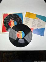 Photo of the vinyl record, album cover, and lyric sleeve laid flat on a white surface, showing front cover artwork and vinyl record label.