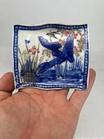 Front view of small rectangular porcelain dish plate held in hand, showing blue and gold border with bird and flower hand-painted design.