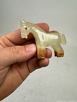 Front and side view of small carved onyx horse figurine held in hand showing translucent white stone with brown accents and detailed carving.