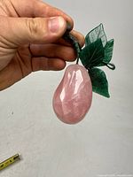 Full view of pear showing rose quartz body with jade green leaves and a dark brown stem held by a hand for scale.