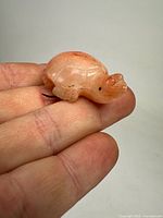 Close-up photo showing the carved turtle stone brooch held between fingers, highlighting the shell detailing and peach-orange color with natural veining.