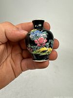 Photo of front and one side of the small famille rose porcelain mini vase held in hand for scale, showing floral patterns in multicolor on black background, vase has no chips or cracks.