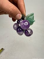 Front hand-held view showing six amethyst grapes with green jade leaves attached to a dark jade vine stem.