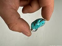 Close-up photo of bright blue turquoise pendant held between fingers showing natural stone patterns and partial silver-tone mounting.