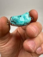 Close-up image of the mounted turquoise pendant held between fingers, showing polished blue-green stone with black veining and silver metal mounting with one loop visible.