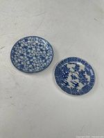 Two small blue and white decorative plates shown side by side on a flat surface. The left plate has dense floral pattern, the right plate has a traditional Japanese scenic design with a geometric border.