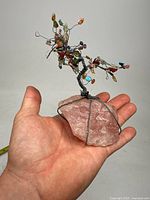 Bonsai tree sculpture held in hand, showing twisted wire branches with multicolored stone pebbles and large rose quartz base.