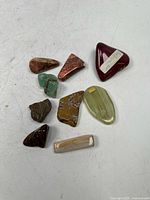 Top down photo of 9 small polished stones of various colors and shapes including red, green, brown, and yellow stones on a white surface.