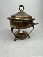 Silver or silver plate footed serving dish on warming stand with lid and ornate handles, photographed from side angle.
