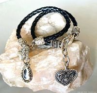 Both black braided bracelets with silver-tone charms displayed on stone