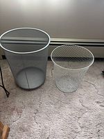 Two medium mesh wastebaskets, one grey and one white, sitting on carpeted floor.