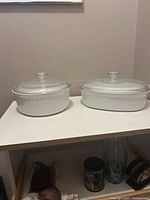 Two white oval casserole dishes with ribbed sides and clear glass lids with round knobs, placed side by side on a white surface.