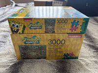 Stacked boxes of SpongeBob SquarePants jigsaw puzzles by Aquarius, showing 1000 piece and 3000 piece counts on side.