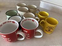 Photo showing assortment of 10 ceramic mugs including 2 red 'KEEP CALM' mugs, 2 yellow 'Cooks Mustard' mugs, and other floral/pale colored mugs