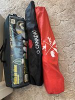 Three carrying bags side-by-side on carpet: the largest bag for a Woods hexagonal dome tent for 4 persons, plus two smaller bags for folding chairs, one black with O Canada and maple leaf, one red with outdoor symbols.