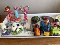 Photo displaying small toy collection including Pink Panther figurines, Snoopy figures, Peanuts character toys, and small colorful toy vehicles arranged on the tray