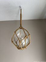 Photo of clear glass fishing float encased in tan rope netting hanging from ceiling hook against beige wall and white curtain background.