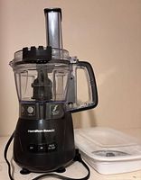 Hamilton Beach 4 cup food processor unit with attached clear plastic container and handle, black base with controls, power cord visible, and white accessory case beside it.