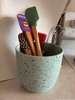 Kitchen utensils including spatulas, brushes, and whisk stored in speckled ceramic holder.