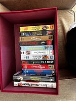 Top-down view of 12 assorted VHS tapes stacked inside a red decorative box. Titles visible include "Black Stallion", "Treasure Island", "Pedrito and the Wolf", "Wind in the Willows", and "James and the Giant Peach." Cases show wear and some labels are faded.