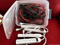 Plastic storage tub filled with at least three white power strips and multiple black and red extension cords. The power strips have multiple outlets and on/off switches. Cords are coiled and tangled.