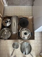 Top view of 8 cookware pieces arranged in a cardboard box, showing pots, pans, and matching glass lids, along with one long narrow pan.