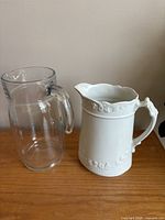 Photo showing clear glass pitcher and white ceramic pitcher side by side, highlighting designs and condition.