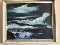 Full view of framed oil painting showing crash of waves on rocky shore with dark sky.