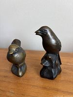 Pair of bronze or brass bird figurines shown together on wooden surface against light wall.