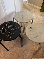 Three metal stools on carpet floor, two gray and one black, showing the perforated tops and tubular legs.