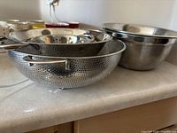 Photo shows five nested stainless steel kitchen bowls and strainers with a reflective finish arranged on a kitchen counter.