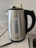Front view of stainless steel electric kettle with black handle, water level indicator visible on side, cord plugged into wall outlet.