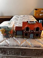 Overall lot showing Fisher-Price playhouse and plastic storage bin containing figures and accessories on mattress surface.
