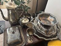 Photo showing an assortment of tarnished silverplate items including tall coffee or tea pot, sugar bowl, covered butter dish, multiple serving trays and goblets.