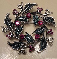 Close-up image of the front side of the brooch showing holly leaves with green rhinestones and red rhinestone berries arranged in a circle.