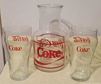 Glass jug in center with two Coca-Cola branded glasses on each side, all clear glass with red text and logos.