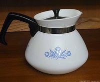 White ceramic Corningware tea kettle with blue Cornflower pattern, black handle and knob, metal band near lid. Shown from side angle.