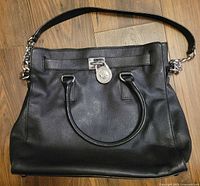 Front view of the black leather satchel bag showing two handles and silver MK lock charm.