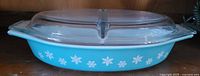 Side view of blue Pyrex oval casserole dish with white snowflakes and clear divided lid, showing the pattern and handles.