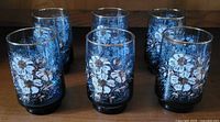 Six blue vintage Libbey St. Clair juice glasses arranged on a wooden surface, showing floral designs with white and gold accents.