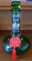 Front view of green glass decanter bottle labeled Captain's Table Canadian Whisky 710 ml with red wax seal pendant around neck and wide base glass stopper.